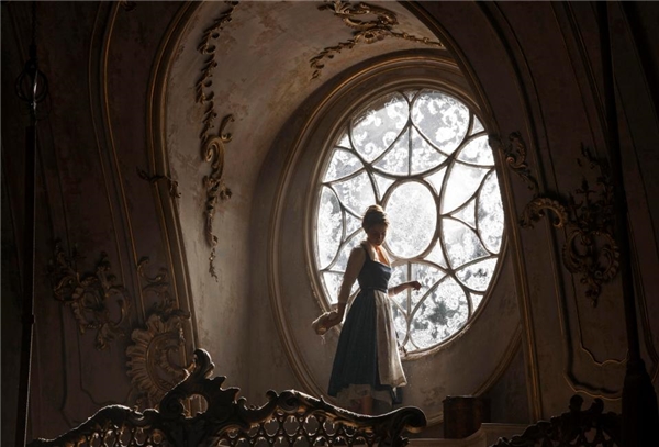 Emma Watson trong vai nàng Belle đứng cạnh khung cửa sổ kính màu lớn trong lâu đài của Quái vật.