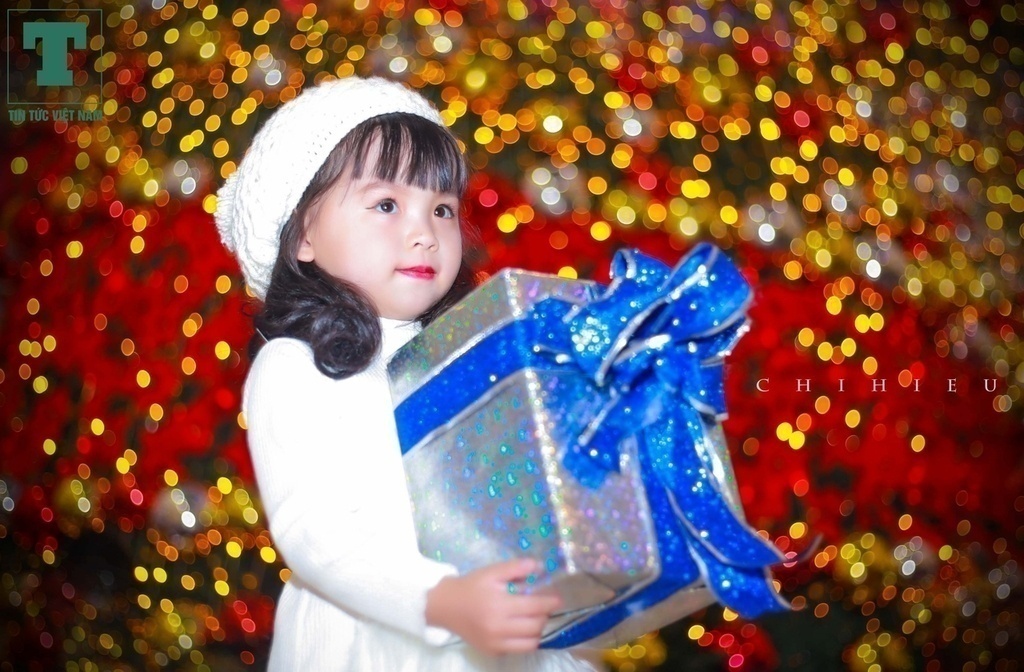 Một em nhỏ thích thú khi cầm món quà lớn đứng chụp hình trước cây thông sáng rực rỡ bởi hàng nghìn ngọn đèn.